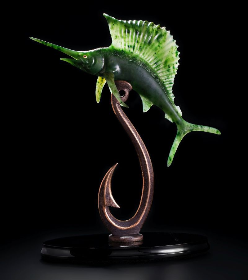 Ein grüner Marlin (Schwertfisch) graviert aus Nephrit, mit Augen aus 999 Gold liegt oben auf einem Haken aus Bronze. alles steht auf einem schwarzen Obsidian Sockel.