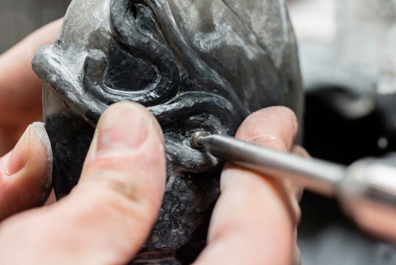 Auf dem Bild sieht man einzelne Finger die einen Stein halten und eine kleine Metallspindel, mit dem dieser Stein graviert wird.