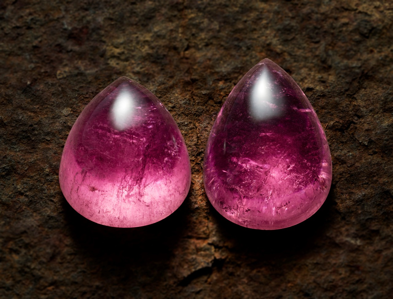 Auf dem Bild sieht man 2 tropfenförmige rosa Turmalin Cabochons auf braunem Untergrund.