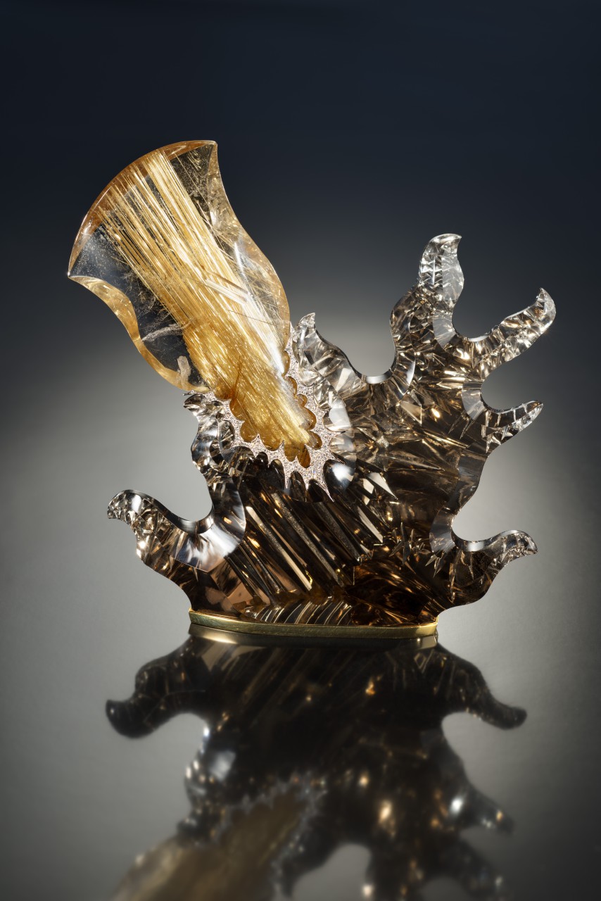 Die Basis dieser Skulptur ist  ein Rauchquarz, geschliffen und graviert  bzw. der viele Einschnitte hat. Die Form erinnert an ein Aufspritzen des Materials bei einem Ausprall, verursacht durch den kometenförmigen Rutilquarz, der von links oben eingefügt ist. Verbunden sind die 2 Teile mit einem gezackten Diamantband, das an Stechpalmen erinnert.